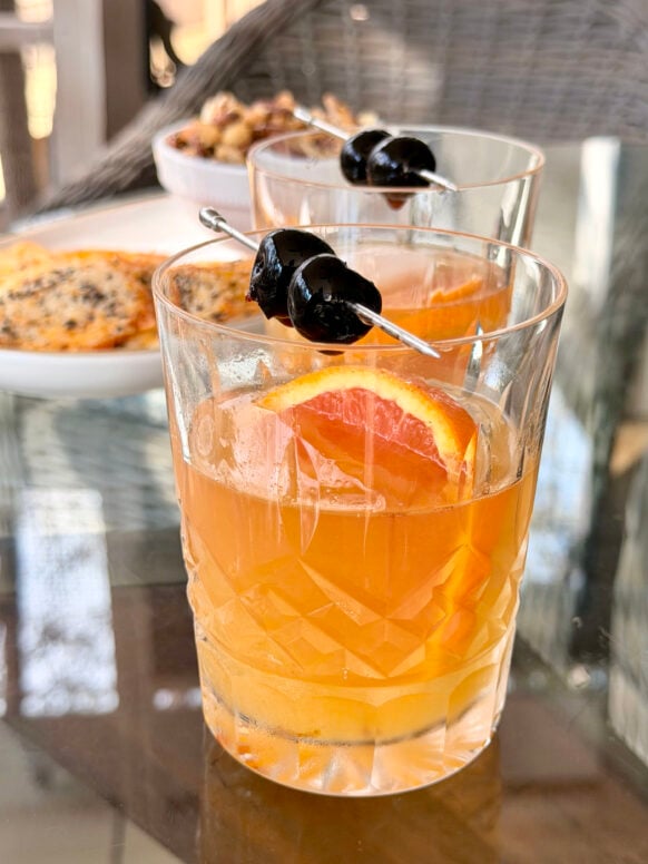 Two blood orange whiskey sours in crystal rocks glasses garnished with blood orange slices and brandied cherries on cocktail picks, cheese crisps visible in the background