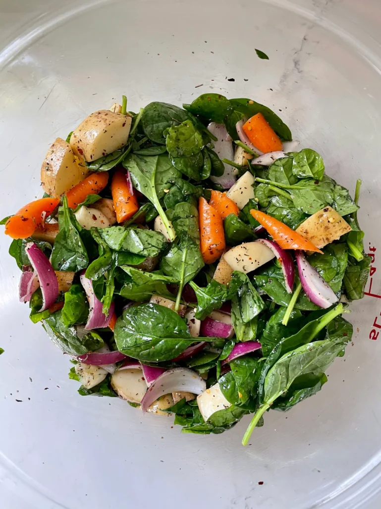 Fresh spinach mixed with seasoned potatoes, carrots, and red onion in a large glass bowl