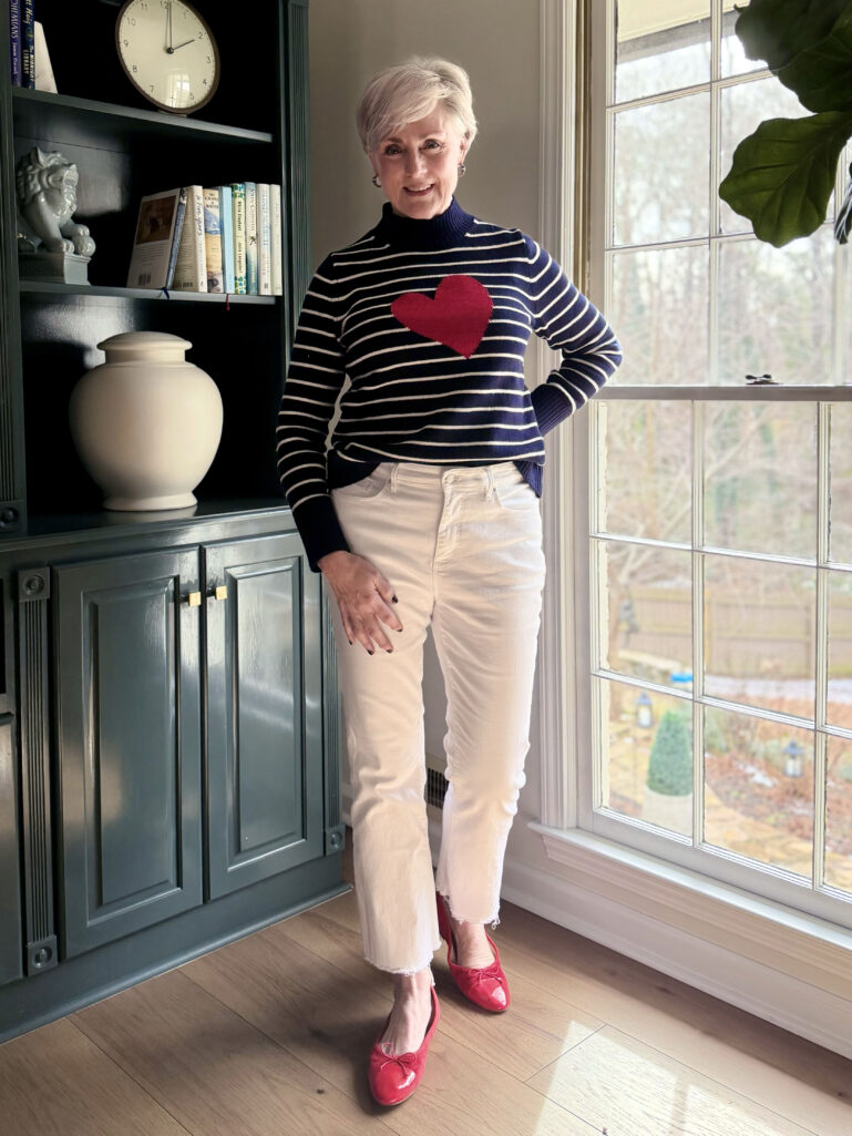 Full-length view of Beth in her heart sweater and cream jeans, hand on hip, winter trees visible through the window