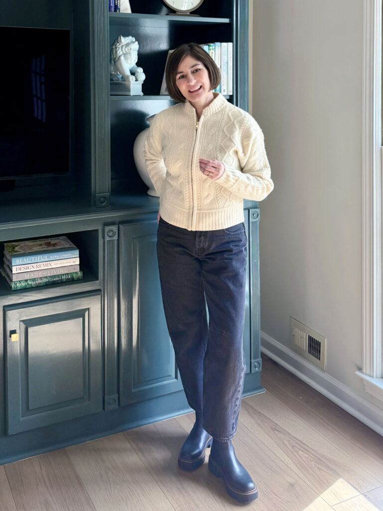 Kelly smiling with hand on hip in her cable knit bomber and washed black jeans, soft winter light from the window