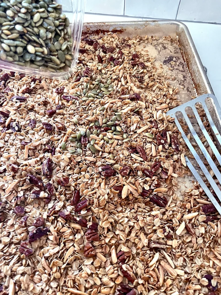 Adding roasted pepitas to golden baked granola on a sheet tray