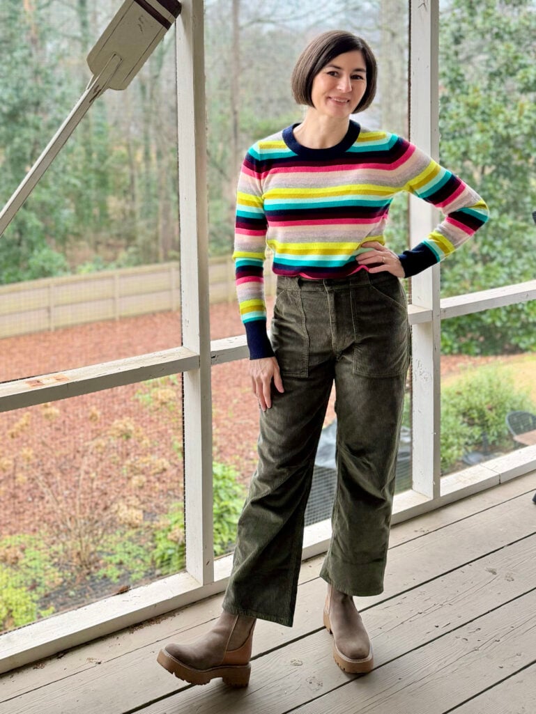 Full-length view of Kelly in her colorful striped sweater and olive corduroys, hand on hip, smiling in the winter light