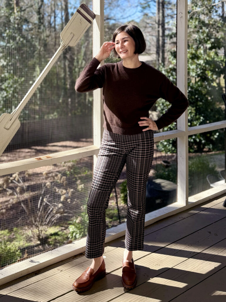 Full-length view of Kelly in her brown sweater and patterned pants, hand brushing hair, smiling in the sunshine
