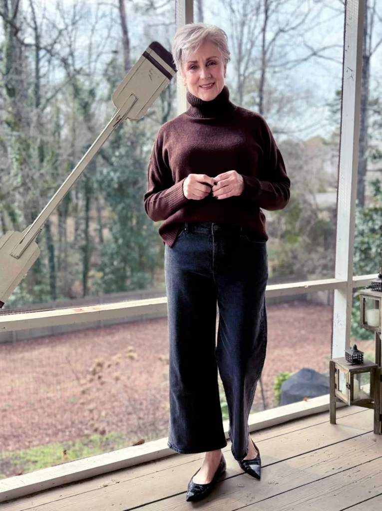 Woman over 50 wearing burgundy oversized turtleneck sweater with wide-leg dark denim jeans and pointed-toe flats