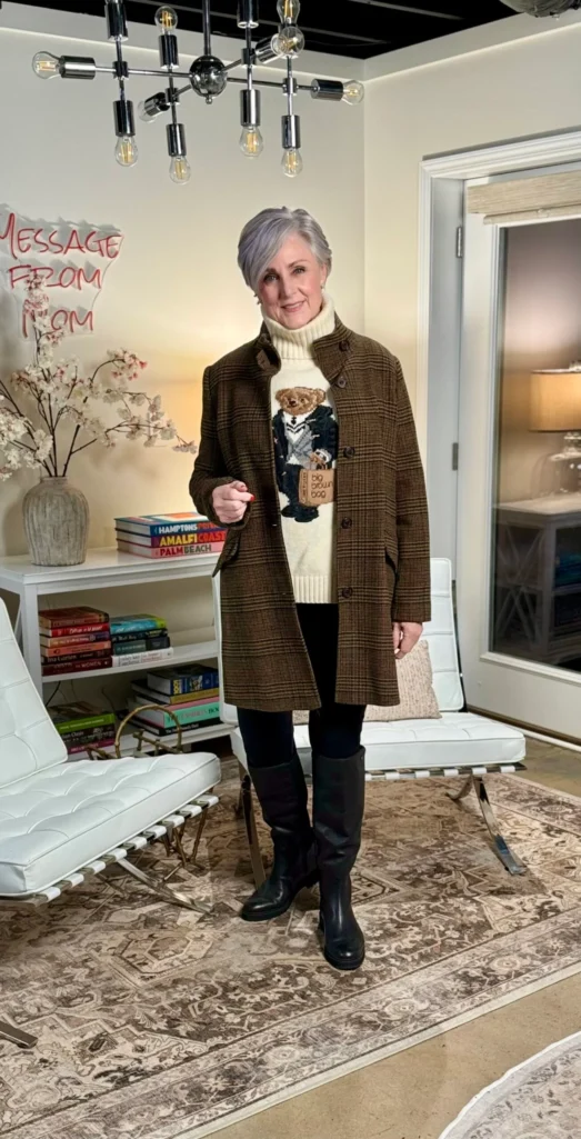 Beth wearing a cream Polo Bear turtleneck sweater under a brown plaid wool coat, with black pants and black knee-high boots — mixing a graphic sweater with a patterned jacket
