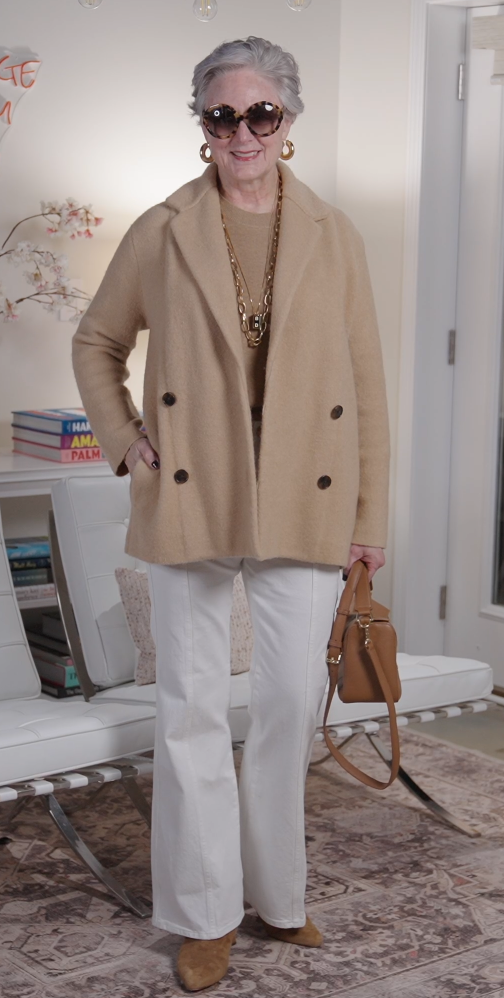 Woman over 50 wearing a camel double-breasted coat with a tan sweater, white jeans, tan ankle boots, layered gold jewelry, and a tan handbag.