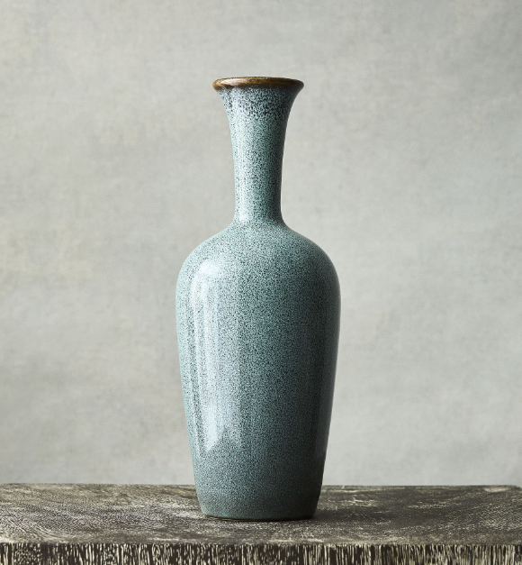 Tall glazed ceramic bud vase in a muted blue-green finish with a narrow neck and rounded base, styled on a wood surface for minimalist home decor.