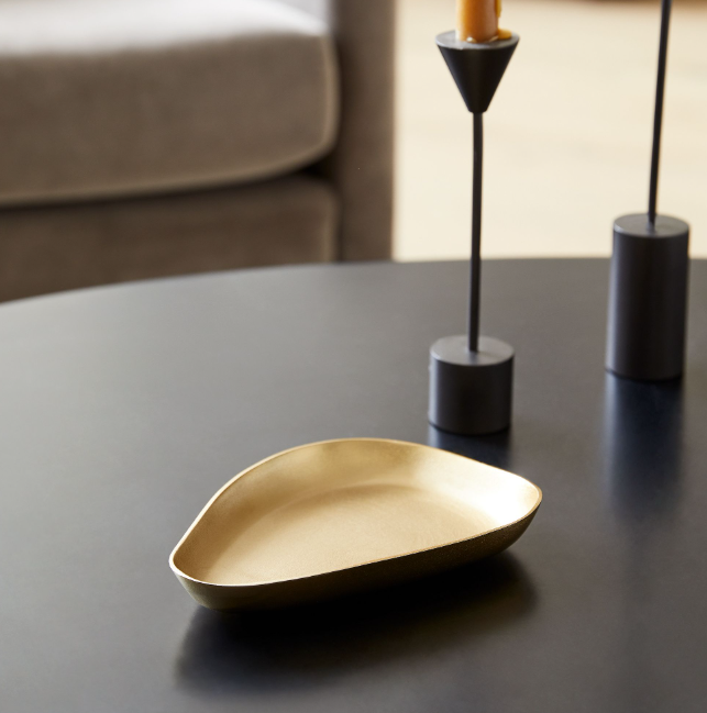 Small organic-shaped metal trinket tray in a warm brass finish, styled on a dark tabletop with modern candleholders for a sculptural accent.