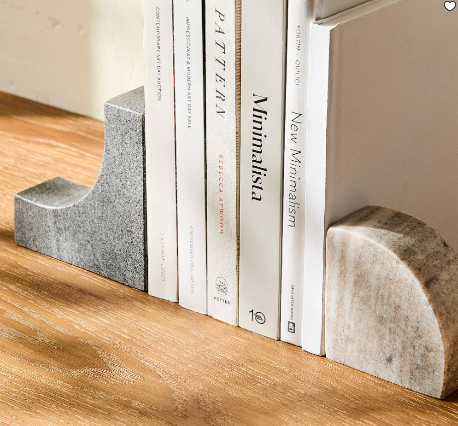 Two-tone marble bookends with curved silhouettes in gray and cream marble, holding neutral hardcover books on a wooden shelf.