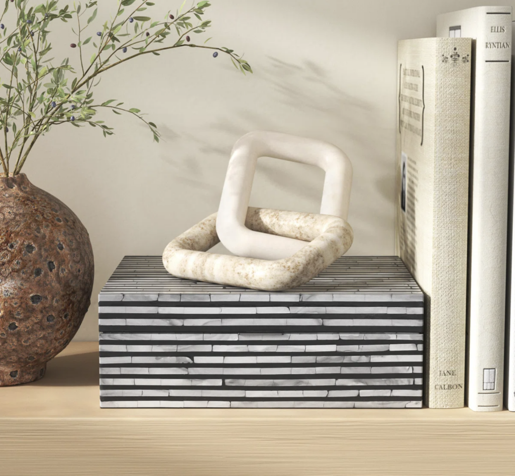 Interlocking chain-link sculpture in neutral stone tones, styled atop a striped decorative box alongside books and a ceramic vase for a modern shelf vignette.