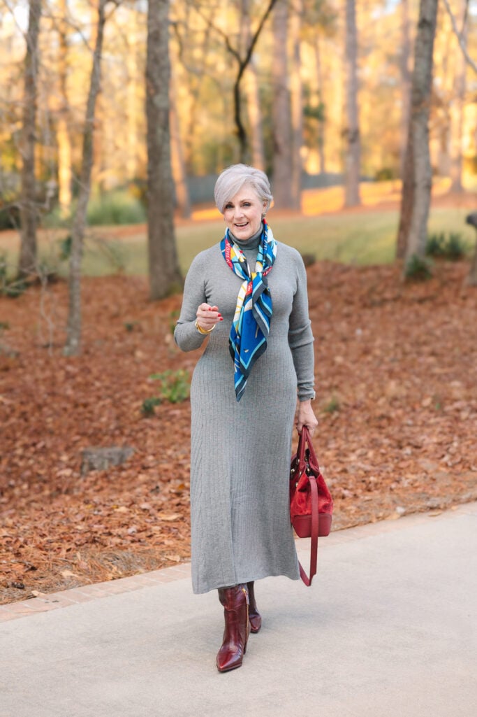 Woman over 50 wearing gray ribbed turtleneck midi sweater dress with a colorful silk scarf and red handbag