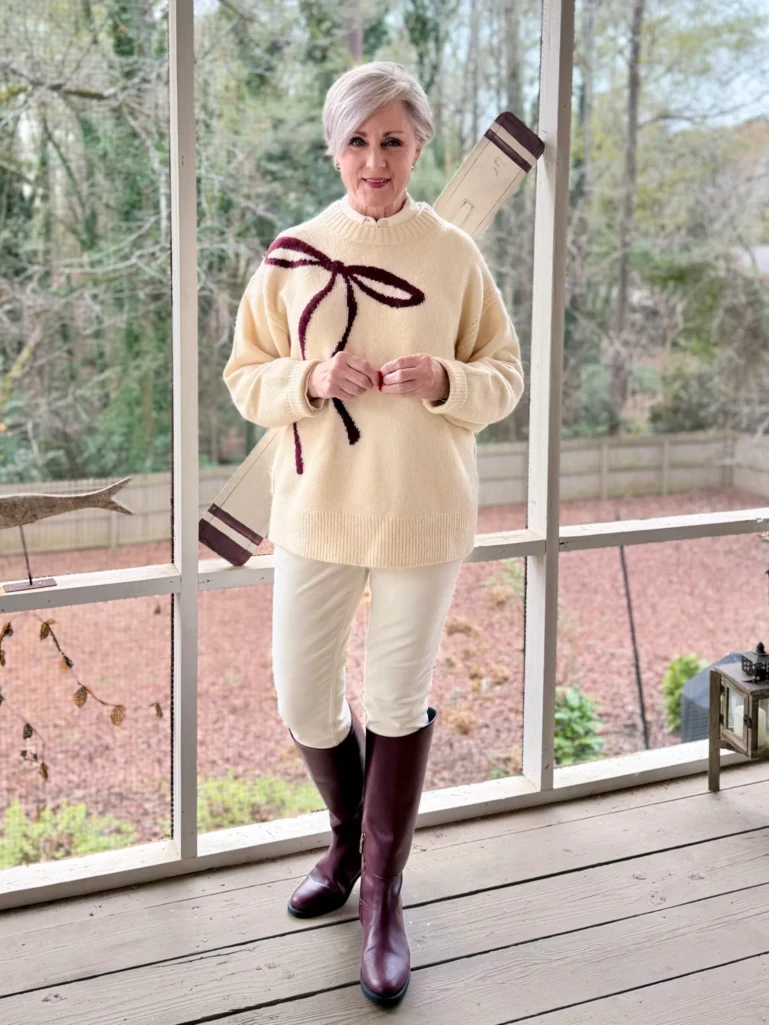 Woman over 50 styling white button-up under cream bow-print sweater with white pants and burgundy riding boots