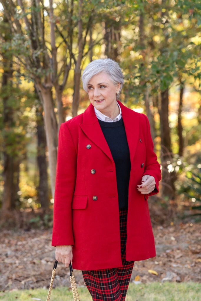 Woman over 50 wearing white button-up under black sweater with red double-breasted coat and plaid pants