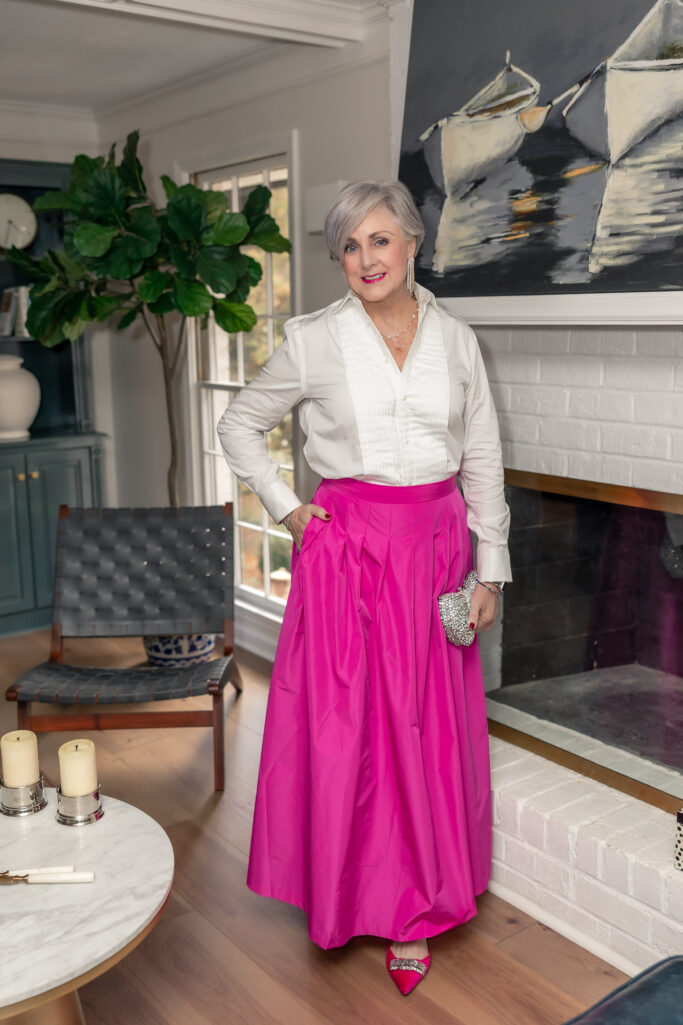 Woman over 50 styling white tuxedo shirt with fuchsia maxi skirt, pink velvet shoes, and crystal clutch
