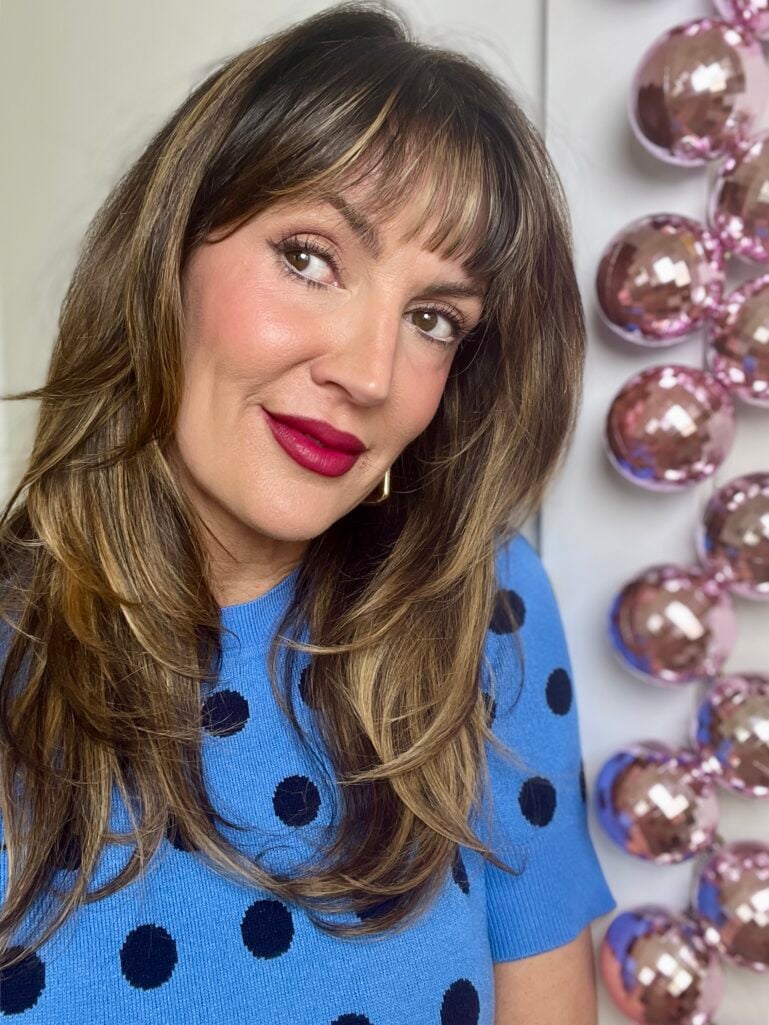 Woman with highlighted brown hair and bangs wearing bright blue polka dot short-sleeve sweater, bold berry matte lipstick, and gold hoop earrings, with pink disco ball decorations in background
