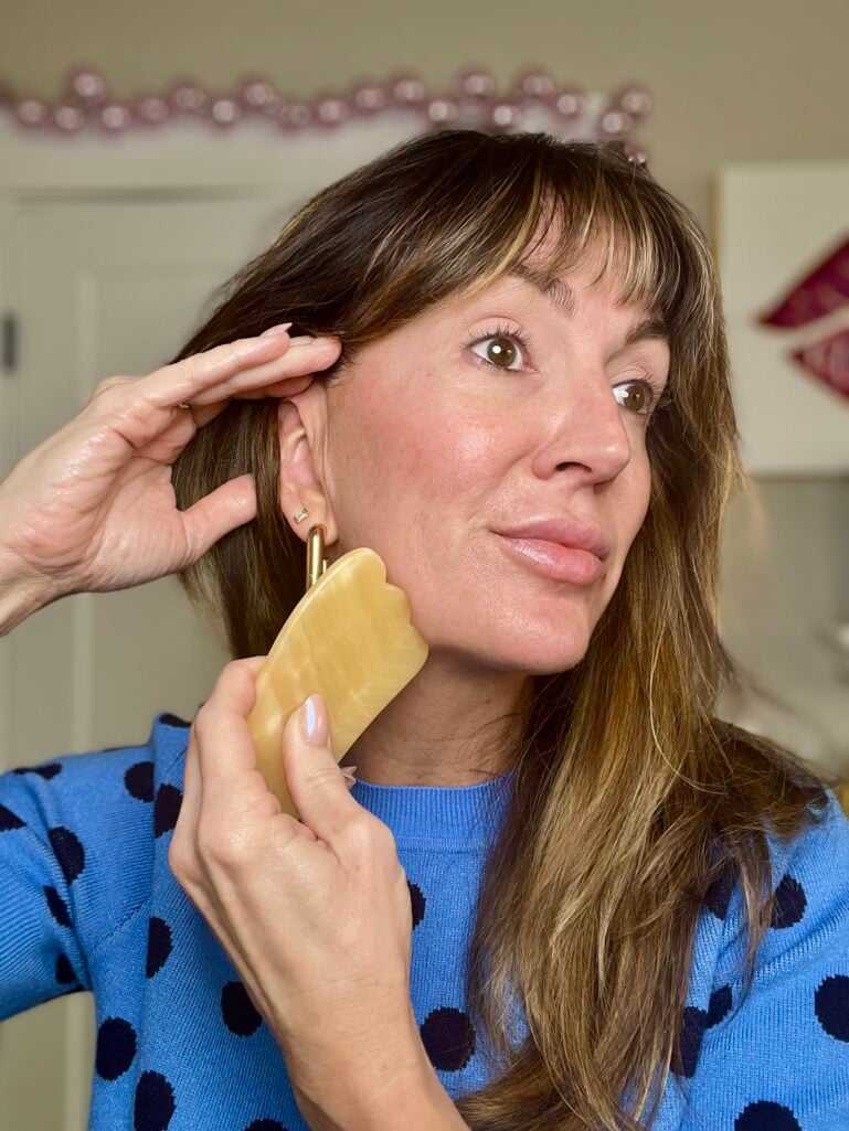 Woman using yellow gua sha stone on jawline in upward motion, wearing blue polka dot sweater and gold geometric earrings, demonstrating facial massage technique with natural bare-faced skin