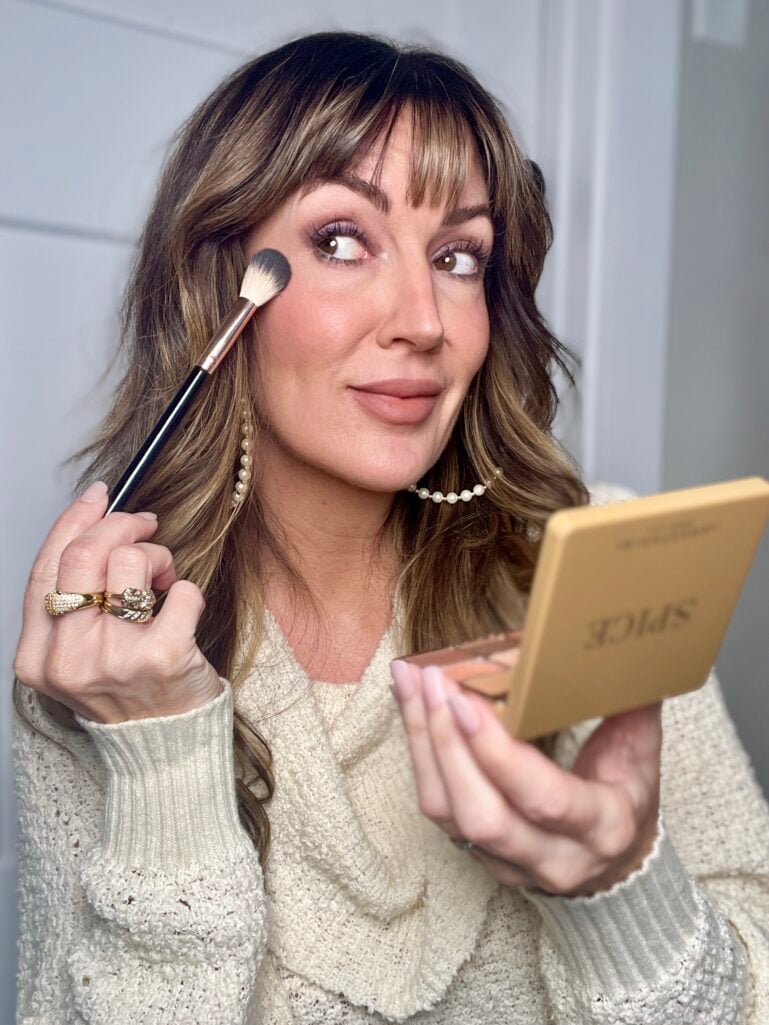 Image 14 (brand_14_.jpg): Woman with highlighted brown hair applying bronzer with black and white contour brush while holding open Anastasia Beverly Hills Spice palette in gold packaging and looking into compact mirror, wearing cream knit v-neck sweater, purple shimmer eyeshadow, rosy cheeks, pearl hoop earrings, and gold stacked rings