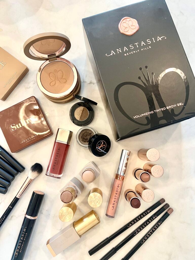 Flatlay of Anastasia Beverly Hills products on marble surface including Sultry eyeshadow palette, bronzer compact, Volumizing Tinted Brow Gel box, brow powder duo, lip gloss, dipbrow pomade, cream eyeshadow singles, brow pencils, lipsticks, and fluffy powder brush