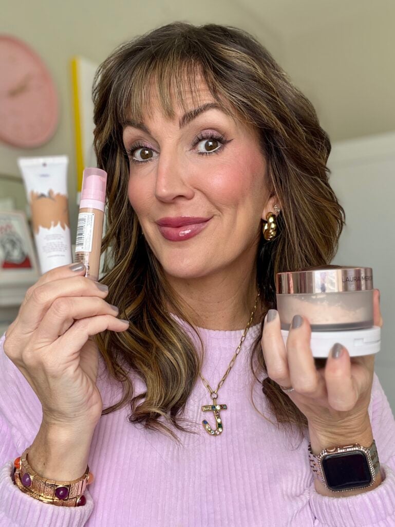 Woman holding NYX Bare With Me concealer serum and Laura Mercier translucent setting powder with Tarte BB Blur visible in background, showing finished makeup look with rosy cheeks, glossy lips, and purple shimmer eyes, wearing lilac sweater, gold earrings, jeweled J necklace, gemstone bracelet, and Apple watch