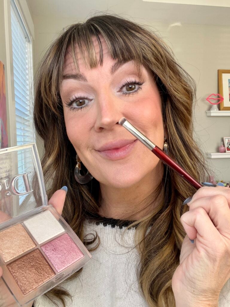 Woman holding Dior Backstage Glow highlighter quad while applying product to bridge of nose with tapered brush, demonstrating nose contouring highlight trick
