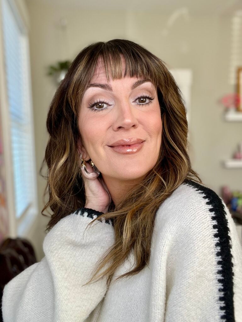 Woman showing finished Pantone Cloud Dancer makeup look with luminous highlighted skin, soft eye makeup, and glossy lips, wearing cream sweater with black chevron stripe detail and smoky quartz hoop earrings