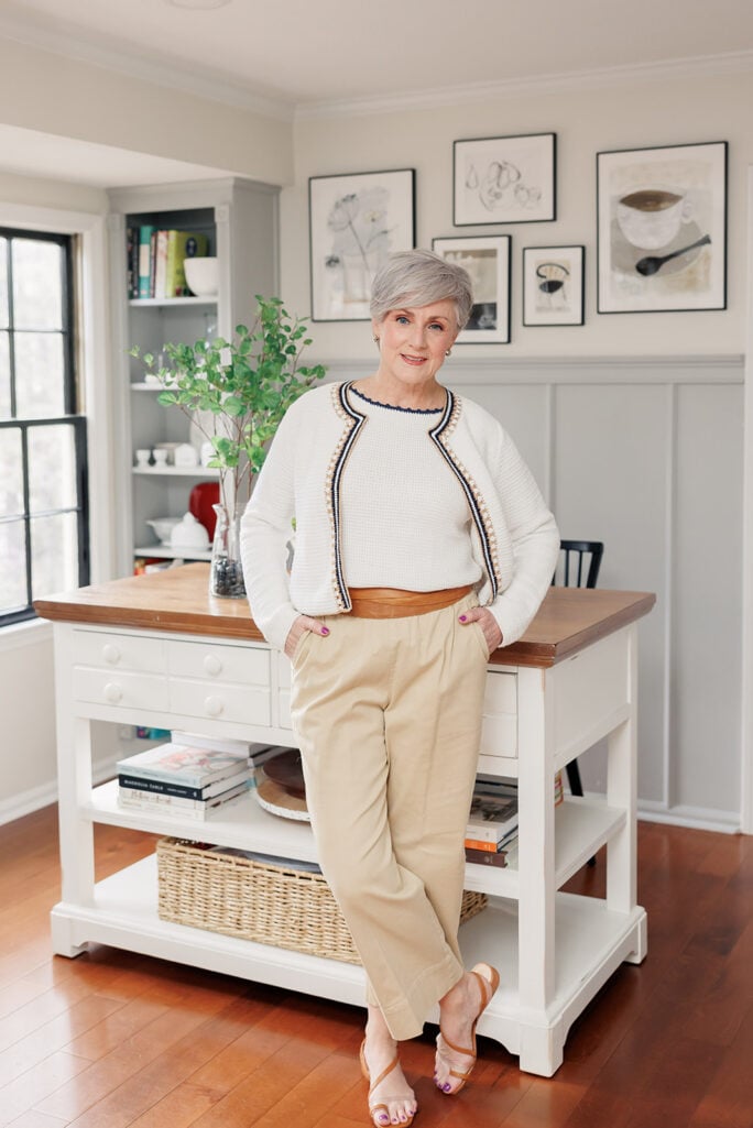 Woman over 50 with silver hair wearing tan pleated wide-leg pants with cognac leather belt, white waffle-knit top, and white open-front cardigan with navy and tan embroidered trim, brown strappy sandals