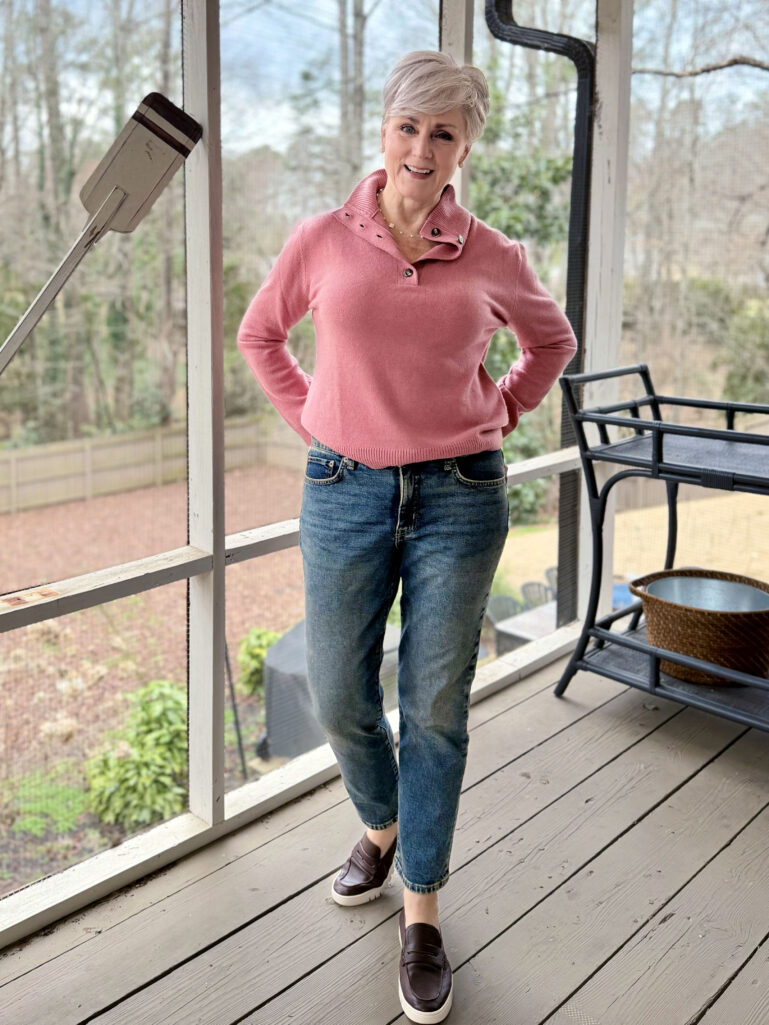 Beth wearing a pink cashmere getaway sweater with button-detail collar, medium-wash straight jeans, and dark brown penny loafers with white soles, hands on hips on the screened porch