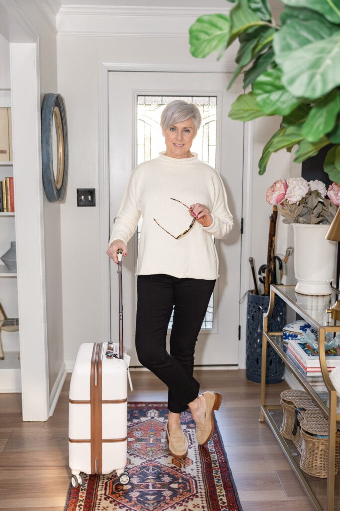 Beth Djalali in a cream cotton rollneck sweater and black ankle pants with suede loafers, holding tortoise sunglasses beside a white rolling suitcase — chic travel outfit for grandmothers on the go.