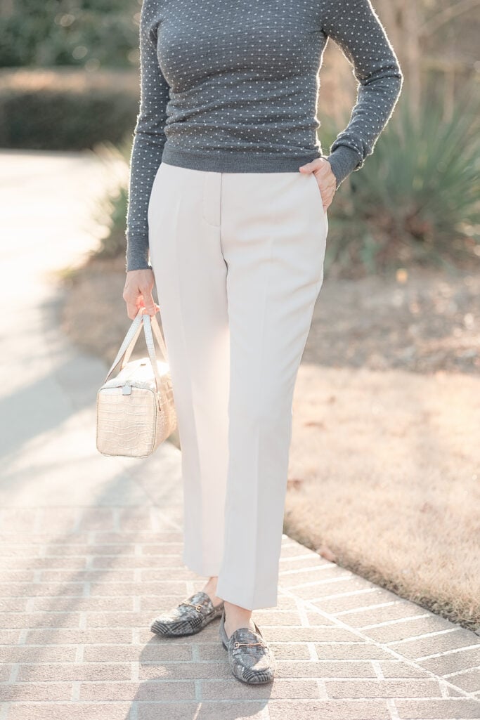 Close-up of charcoal polka dot fitted crewneck sweater tucked into cream Quince stretch crepe ankle pants with plaid sequin loafers and gold croc-embossed bag — showing pant drape and ankle length on a petite frame