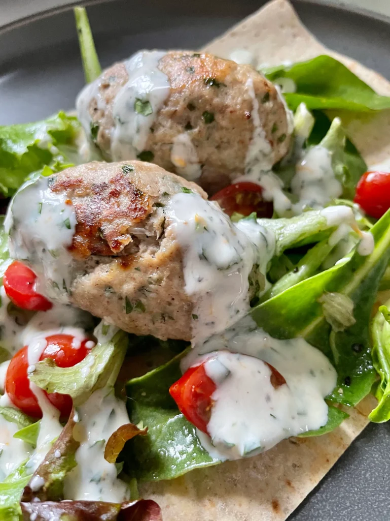 Two golden chicken kofta nestled in spring greens and halved grape tomatoes with yogurt sauce on whole-grain lavash