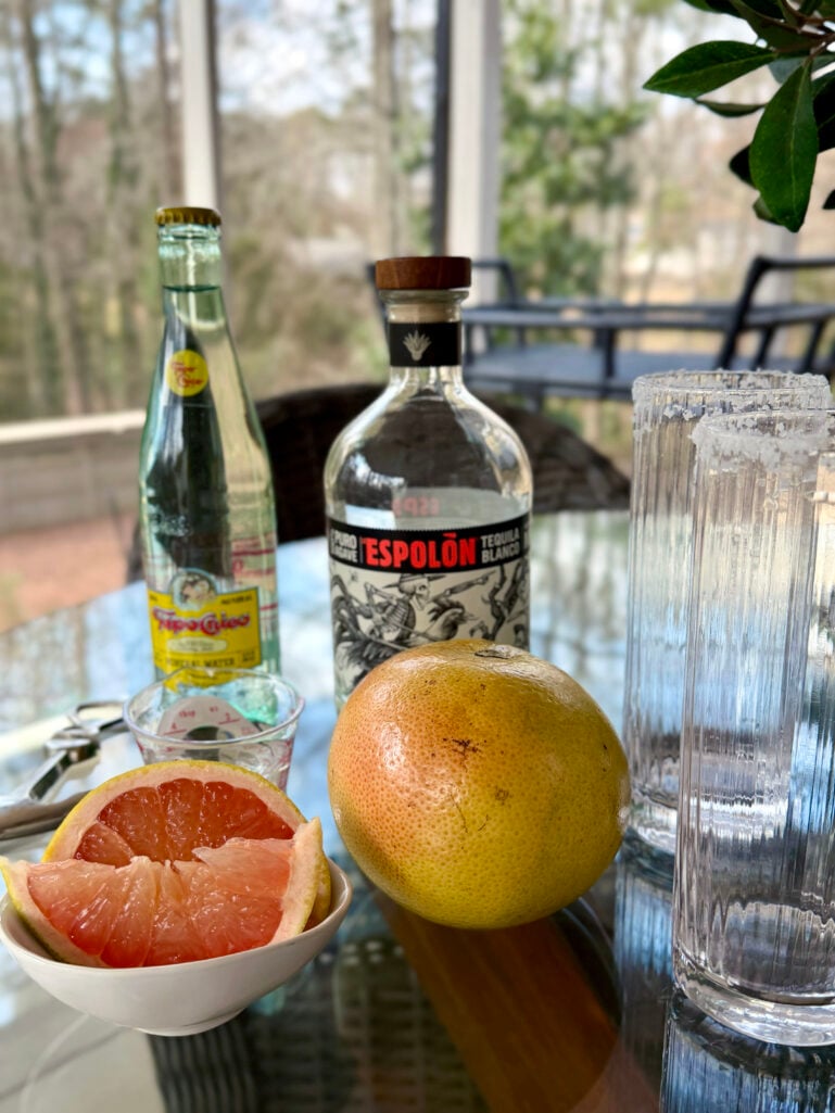 Grapefruit Paloma ingredients on a glass table: Espolòn blanco tequila, Topo Chico sparkling water, fresh grapefruit halves, a measuring glass, and ribbed highball glasses on the screened porch