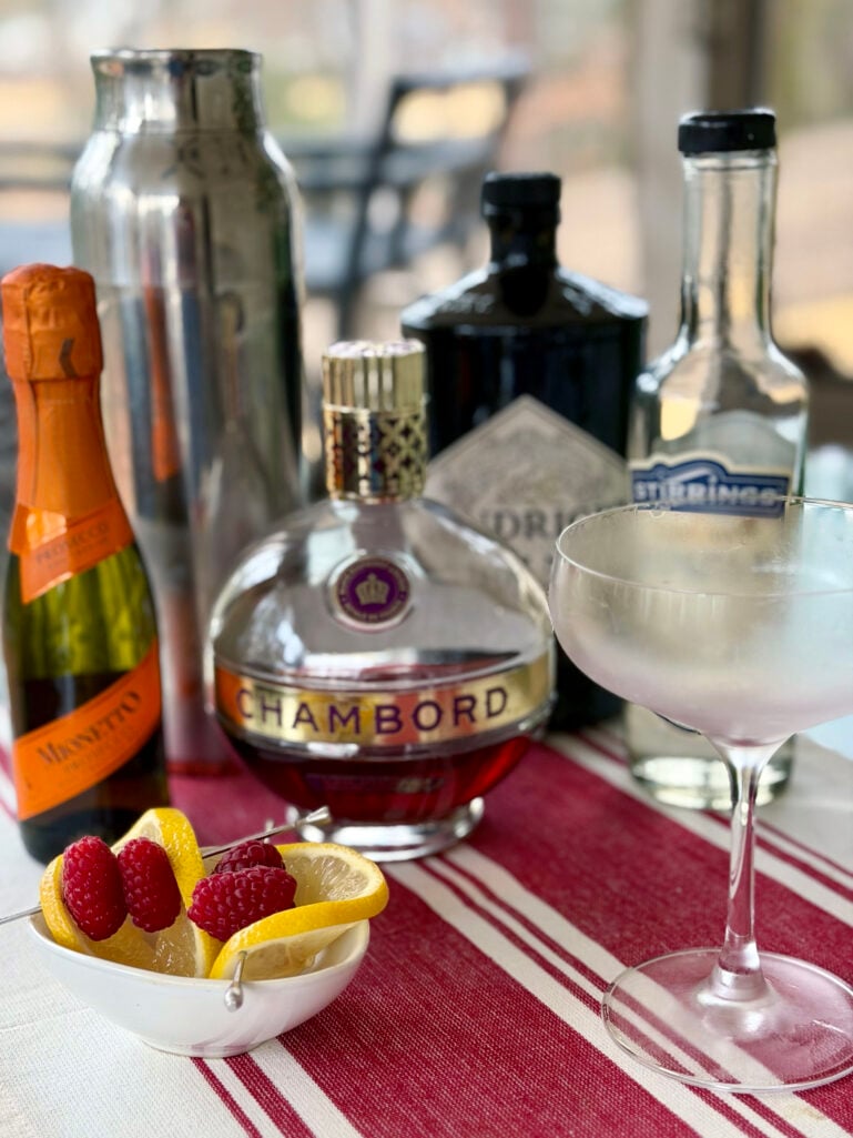 Raspberry French 75 ingredients arranged on a red striped linen: Mionetto Prosecco, Chambord, gin, Stirrings simple syrup, a cocktail shaker, fresh raspberries and lemon slices, and a blush-pink cocktail in a coupe glass