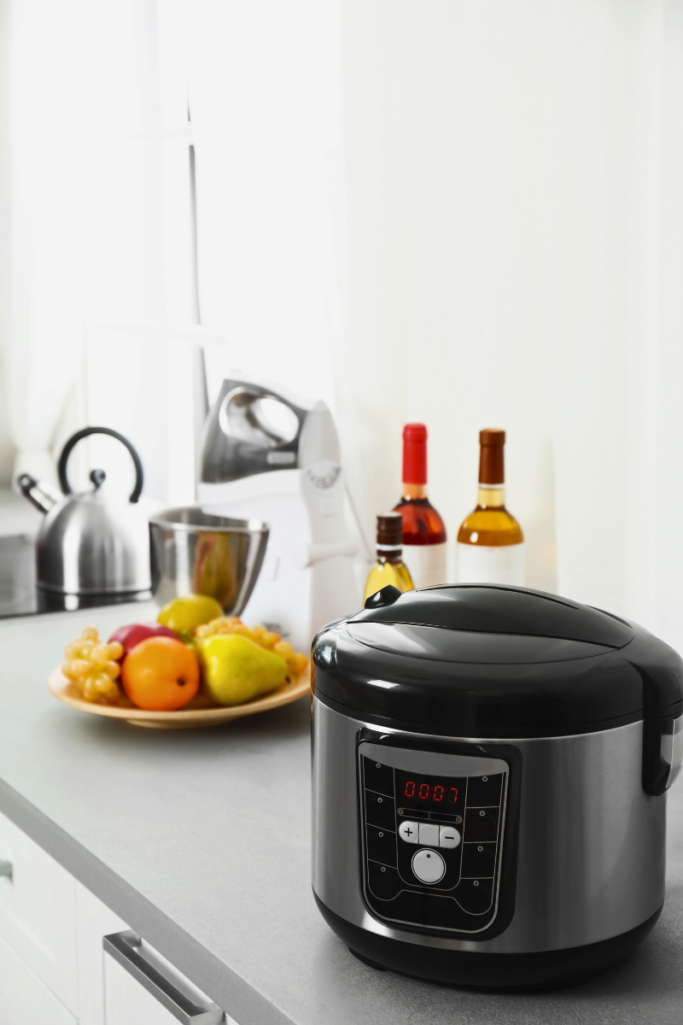 Modern kitchen countertop with a sleek black electric pressure cooker in the foreground, digital display illuminated, with a bowl of fresh fruit, wine bottles, and stainless steel appliances softly lit by natural light in the background.