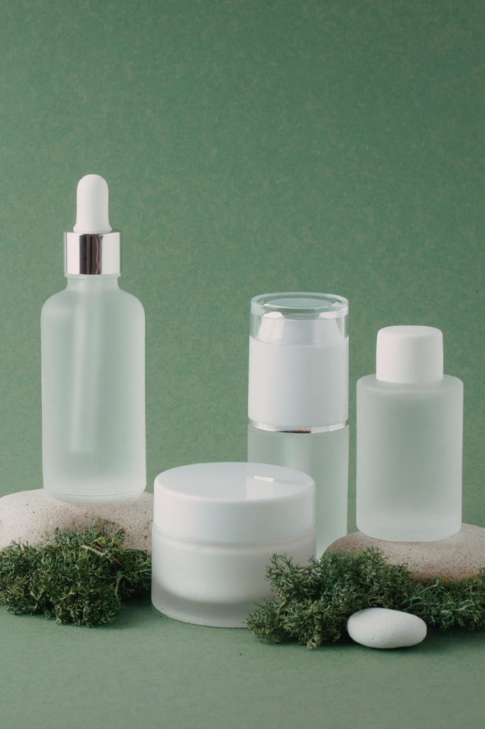 Minimalist skincare and makeup bottles in frosted glass packaging arranged on stone blocks with green moss accents against a muted sage green background.