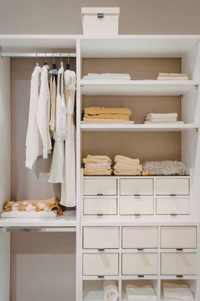 Organized white closet with built-in shelves and drawers, neutral-toned clothing on hangers, and neatly folded cream and mustard towels stacked for a clean, minimalist look.