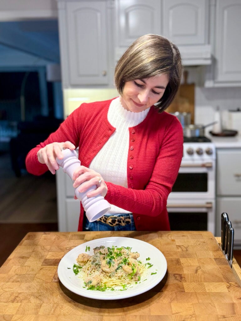 Kelly in her red cardigan grinding fresh pepper over a plate of Creamy Garlic Butter Shrimp served over spaghetti