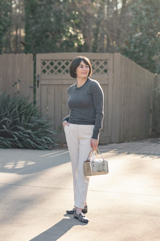 Kelly standing with hand in pocket wearing charcoal polka dot crewneck sweater and cream stretch crepe ankle pants with plaid sequin bit loafers and gold bag — relaxed petite transitional styling