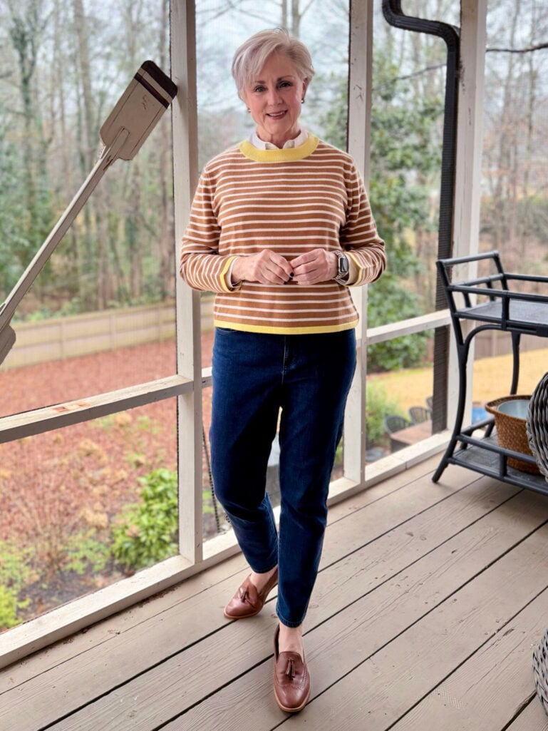 Woman over 50 in a camel and cream horizontal striped sweater with yellow trim layered over a white button-down collar, styled with slim ankle jeans and cognac tassel loafers for a preppy casual look.