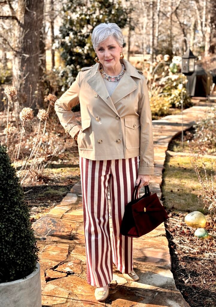 Beth Djalali wearing a khaki cropped trench coat over a gray top, styled with burgundy and cream striped wide-leg pants, cream flats, and a burgundy velvet bag