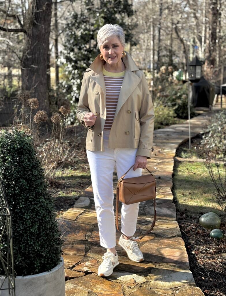 Beth Djalali wearing a khaki cropped trench coat with gold buttons over a striped tee, styled with white jeans, white sneakers, and a cognac leather crossbody bag