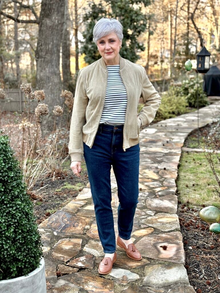 Beth Djalali wearing a cropped sand-colored suede bomber jacket with a navy and white striped tee, dark straight-leg jeans, and cognac tassel loafers