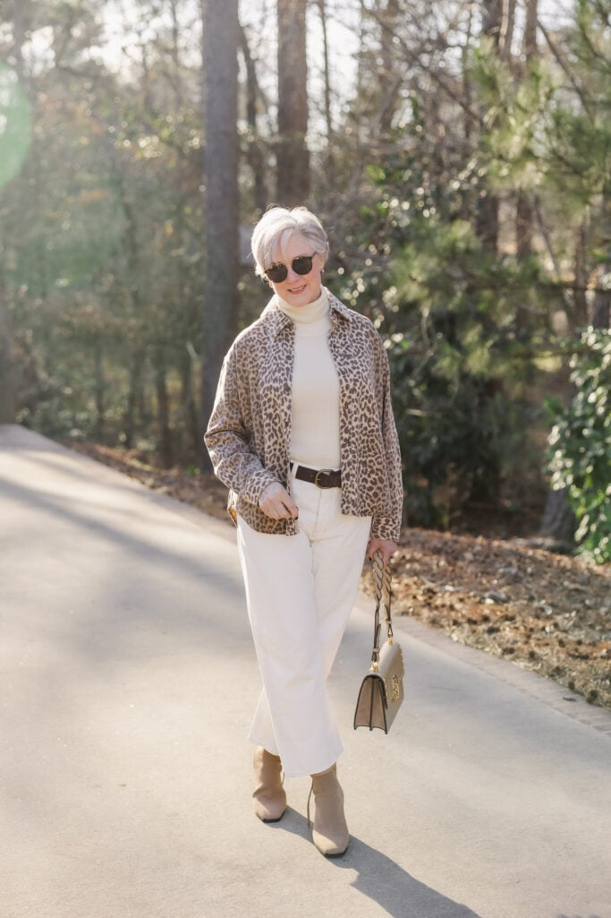 Woman over 50 with silver hair wearing leopard print shirt jacket over ivory turtleneck and cream barrel jeans with tan ankle boots, dark belt, neutral shoulder bag, and tortoiseshell sunglasses