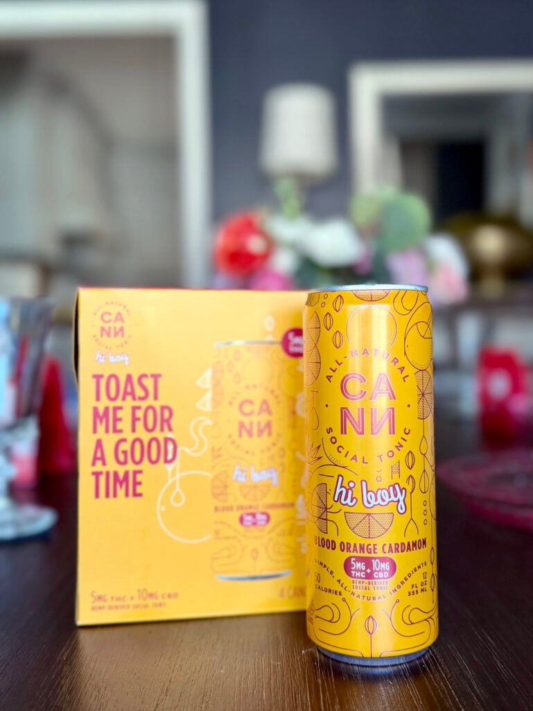 Cann Hi Boy Blood Orange Cardamom social tonic can and four-pack box on a dark wood table, with Valentine's flowers blurred in the background