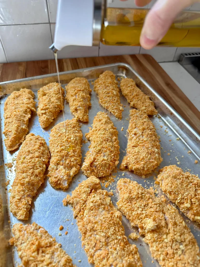 Drizzling olive oil over Cheez-It breaded chicken tenders on a sheet pan before baking