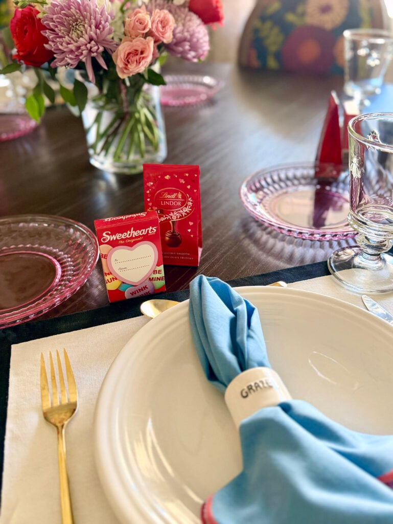 Valentine's Day place setting with a white dinner plate, blue linen napkin with ceramic napkin ring, gold fork, pink depression glass plate, Sweethearts candy box, Lindt Lindor truffles, and a pink and red floral centerpiece