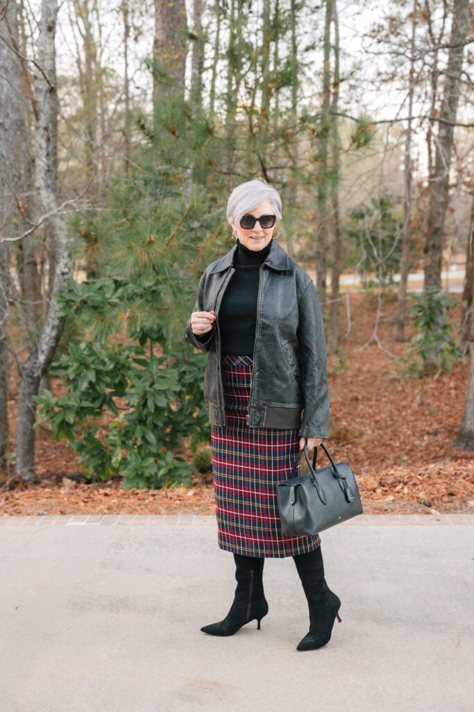 Beth wearing a red tartan plaid midi skirt with a black ribbed turtleneck, black leather bomber jacket, black suede pointed-toe boots, and a black leather satchel — incorporating red into a print for women over 50