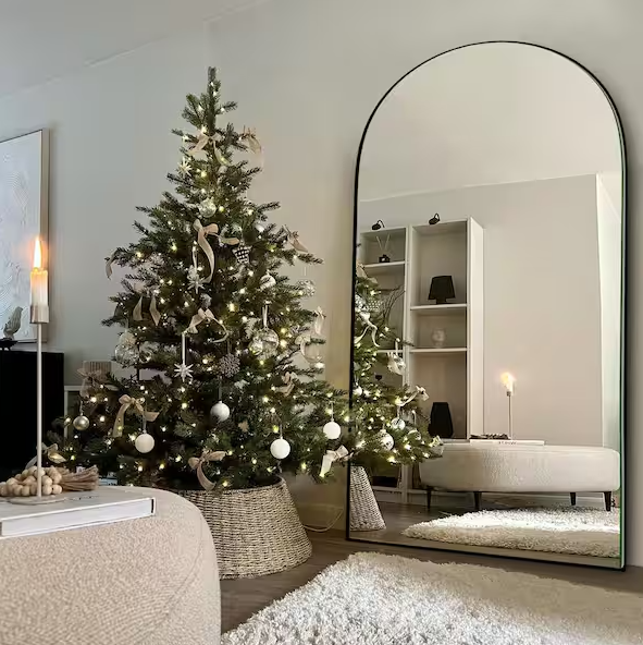 Arched floor mirror leaning against a wall, reflecting a softly lit living room with a decorated Christmas tree, neutral furnishings, open shelving, and a cozy white rug for a warm, minimalist holiday feel.