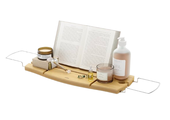 Umbra Aquala expandable bamboo bathtub caddy with stainless steel handles, styled with open book, candle, bath oil, and soap dispenser for a spa-like soak.