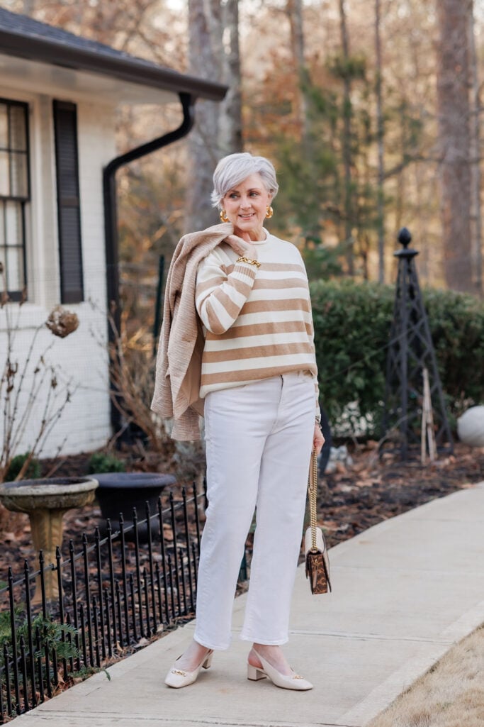 Woman over 50 with silver hair wearing camel and ivory striped crewneck sweater with white straight-leg jeans, oatmeal knit jacket draped over shoulders, nude patent slingback heels with gold horsebit detail, gold hoop earrings, and leopard print crossbody bag