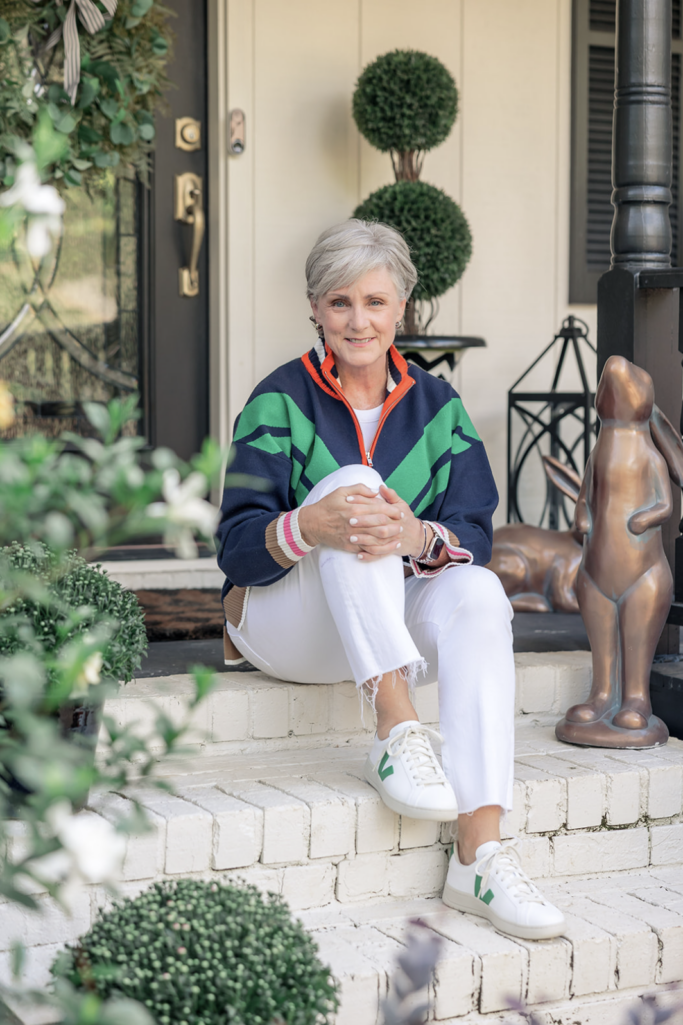 Woman over 50 sitting on front porch steps wearing navy and green colorblock quarter-zip sweater with white raw-hem jeans and white Veja sneakers with green logo